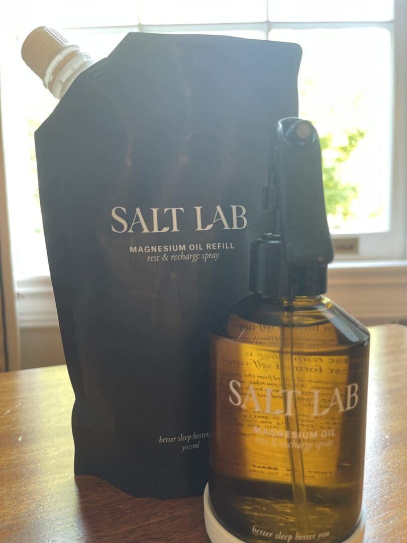 Salt Lab magnesium oil spray and refill pouch on a wooden table near a window, used for relaxation, muscle recovery, and better sleep.