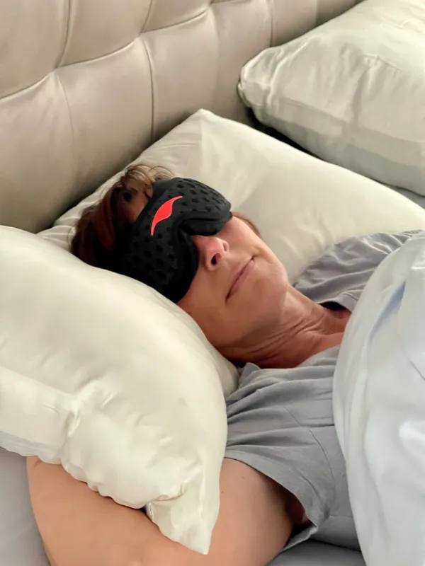 Person resting with an eye mask on a bed, surrounded by pillows.