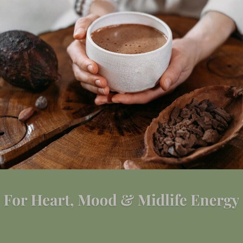Hands holding a cup of cacao on a wooden table with cacao nibs — representing heart, mood, and midlife energy benefits.