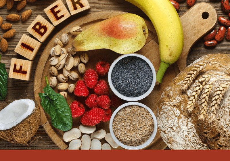 High-fiber foods including bananas, pears, raspberries, oats, nuts, and seeds on a wooden board with the word FIBER spelled in blocks. Text overlay: How much do we really need?